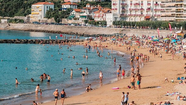 Una playa concurrida, gente nadando y tomando el sol, rodeada de edificios de varios pisos con balcones.