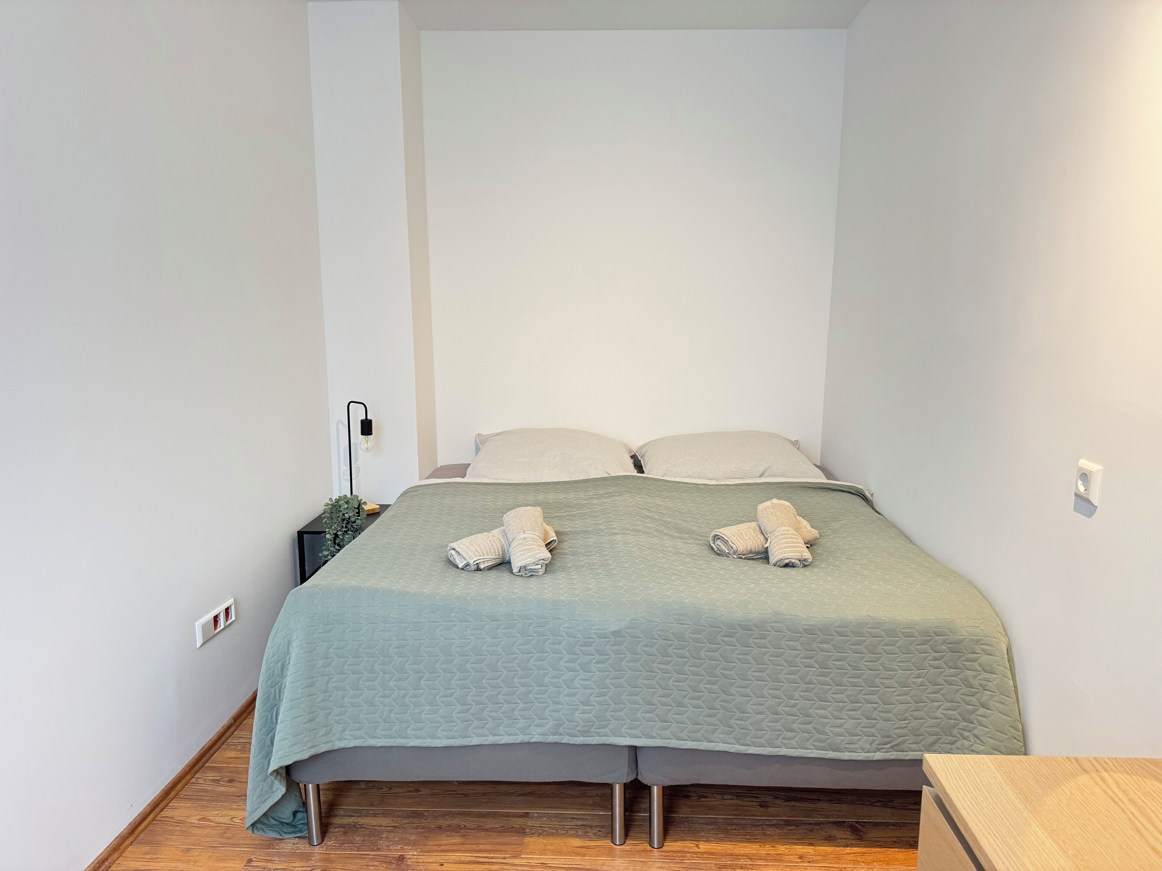 Un lit bien fait, recouvert d’une couette verte, avec deux serviettes enroulées et une table de chevet surmontée d’une lampe.