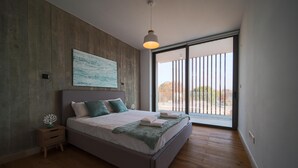 Une chambre avec un grand lit, une table de chevet, une lampe fixée au mur et une porte-fenêtre coulissante donnant sur l’extérieur.