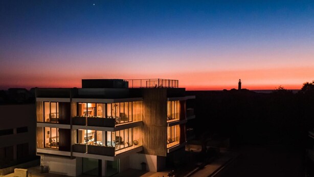A multi-story building with illuminated interior spaces, a balcony, and a clear view of the surrounding area at dusk. - Avenue 202 1 BED - One Bedroom Apartment, Sleeps 2 (Pafos)