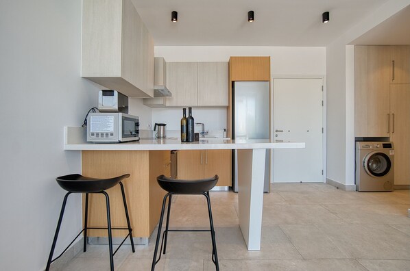 A modern kitchen with a white countertop, two black bar stools, a microwave, a coffee maker, and a washing machine in the background.