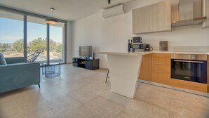 A modern kitchen with a built-in oven, microwave, and coffee maker. There is a dining area with a sofa and a TV. The room has large windows offering a view of the outdoors. - Avenue 202 1 BED - One Bedroom Apartment, Sleeps 2 (Pafos)