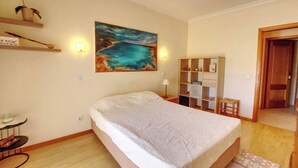 A bedroom with a bed, a painting, a nightstand, a lamp, a shelf with items, and a door. - Vacation Apartment In Portimao (Portimão)
