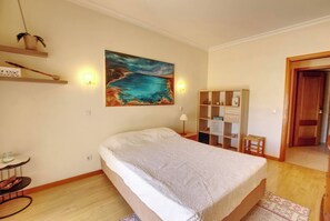 Una camera da letto con un letto, un quadro, un comodino, una lampada, uno scaffale con vari oggetti e una porta.