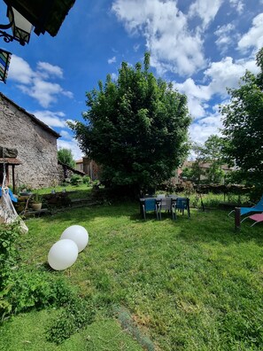 Une cour arrière verdoyante avec un bâtiment en pierre, un grand arbre et du mobilier d’extérieur.