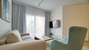 Un salon comprenant un canapé, un fauteuil, une table basse, un téléviseur et une fenêtre avec des rideaux.