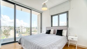 Une chambre avec un grand lit, une table de chevet et une fenêtre donnant sur des immeubles et de la verdure.