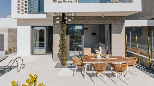 Een modern, tweelaags huis met een patio, een houten tafel en rieten stoelen, een glazen toegangsdeur en uitzicht op het omliggende landschap.