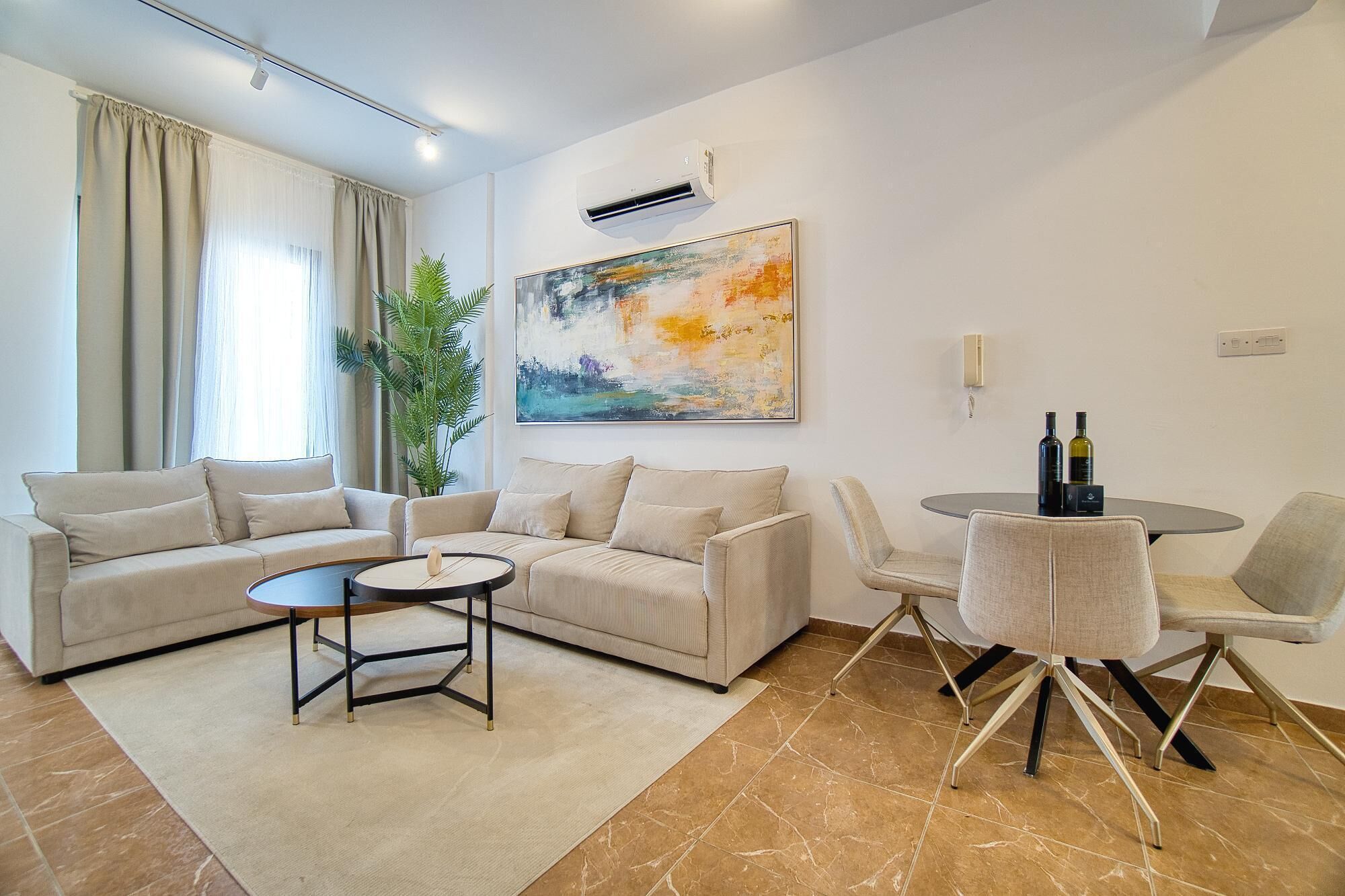 A modern living room with a beige sofa set, a round coffee table, a dining table with chairs, and a large abstract painting on the wall.