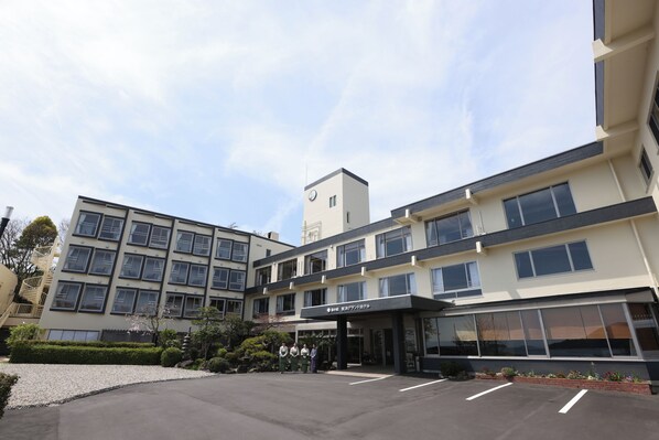 A multi-story hotel with a modern design, featuring a central entrance, a clock tower, and a parking area with a gravel driveway. - Miyahama Grand Hotel (Hatsukaichi)