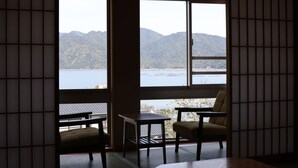 A room with tatami flooring, a low table, two chairs, and a view of a lake and mountains through large windows. - Miyahama Grand Hotel (Hatsukaichi)
