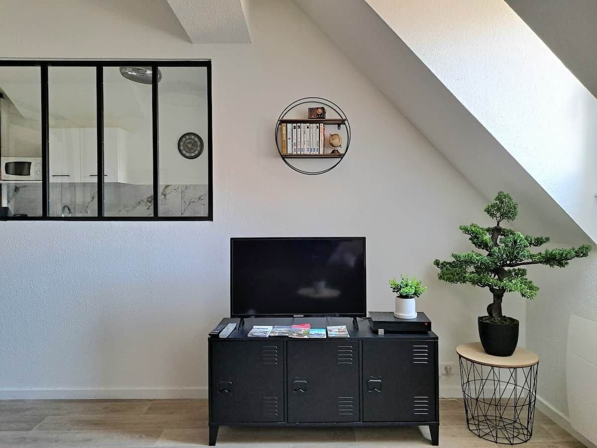 Ein Wohnzimmer mit einem Flachbildfernseher auf einem schwarzen Schrank, einem kleinen runden Regal mit Büchern und einer Uhr, einem Bonsai-Baum auf einem runden Tisch und einer durch eine Glastür sichtbaren Küche.