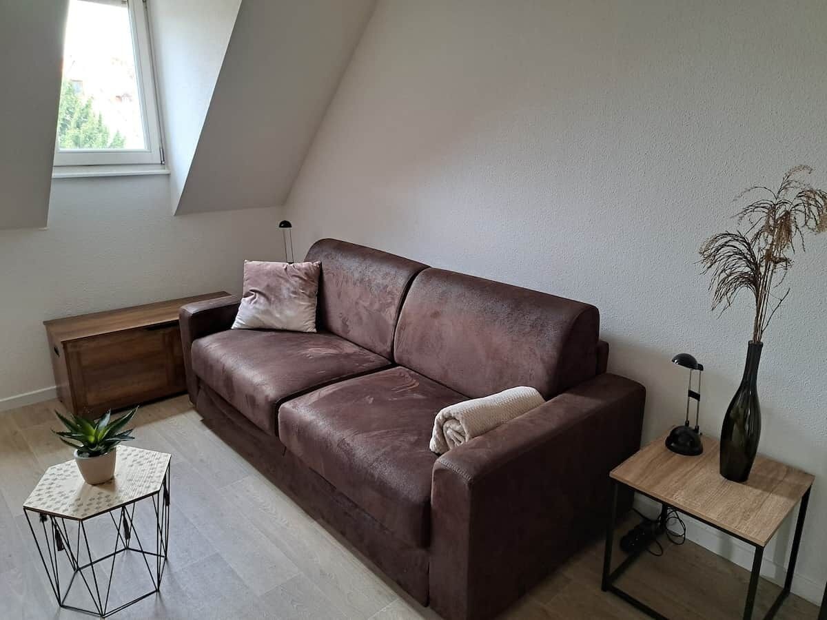 A modern living room with a brown leather sofa, a small side table with a lamp, a potted plant, and a wooden bench.