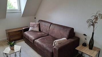 A modern living room with a brown leather sofa, a small side table with a lamp, a potted plant, and a wooden bench.