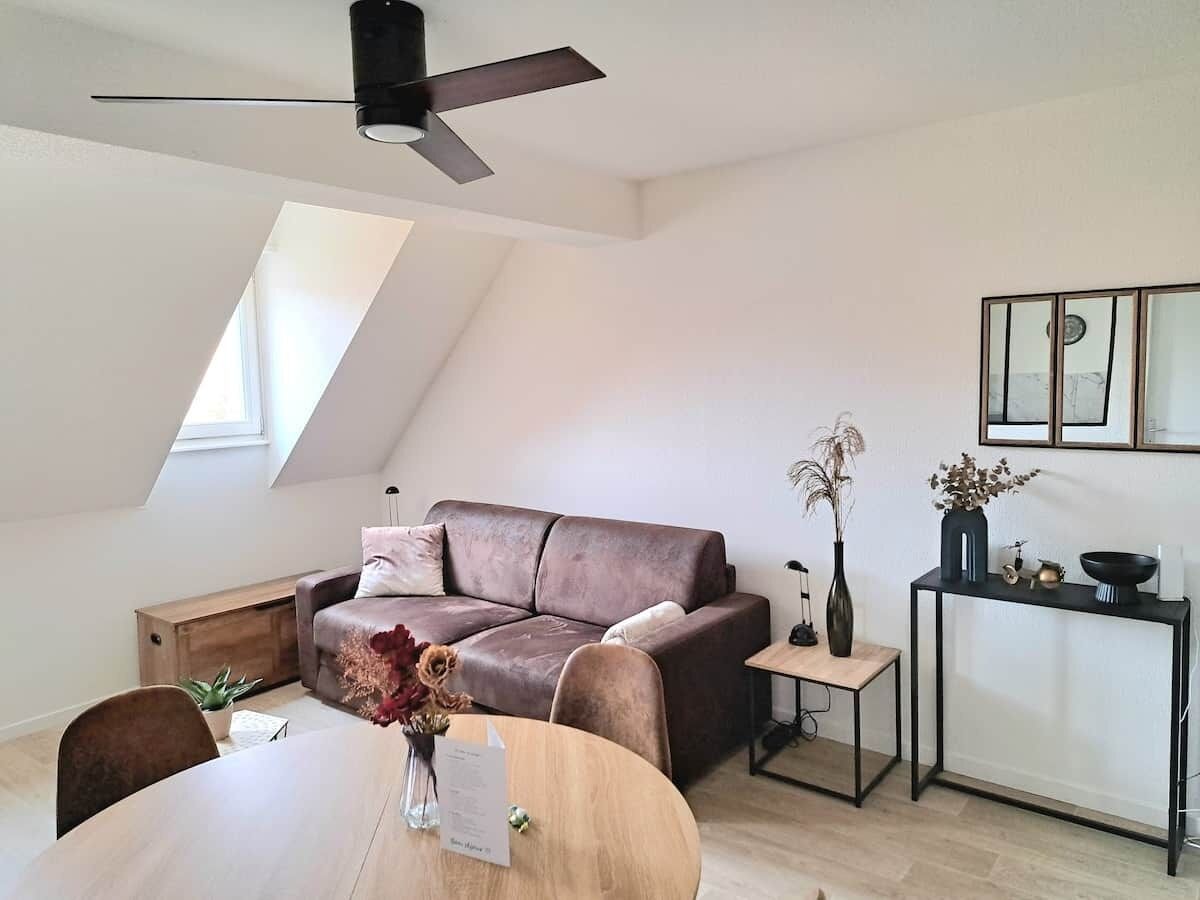 Ein modernes Wohnzimmer mit einem braunen Ledersofa, einem Esstisch mit Blumenvase, einem kleinen Beistelltisch und einem Deckenventilator.