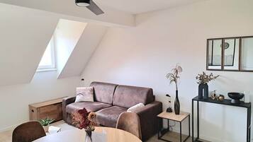 A modern living room with a brown leather sofa, a dining table with a vase of flowers, a small side table, and a ceiling fan.