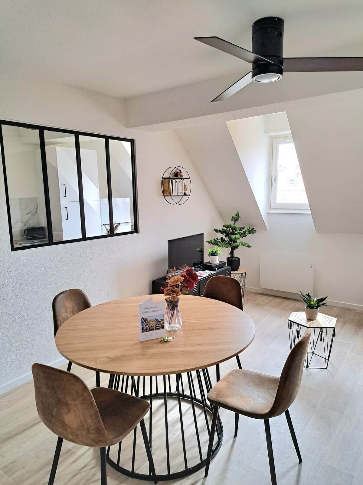 A modern dining area with a round table, four chairs, a ceiling fan, a large mirror, and a potted plant.