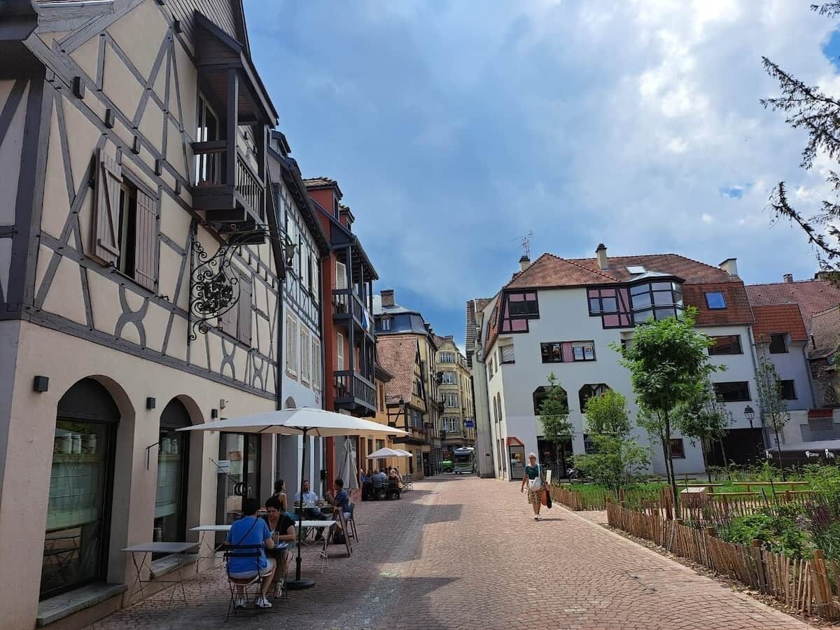 Una calle adoquinada flanqueada por edificios tradicionales de entramado de madera, con mesas de café al aire libre y un paseo peatonal.