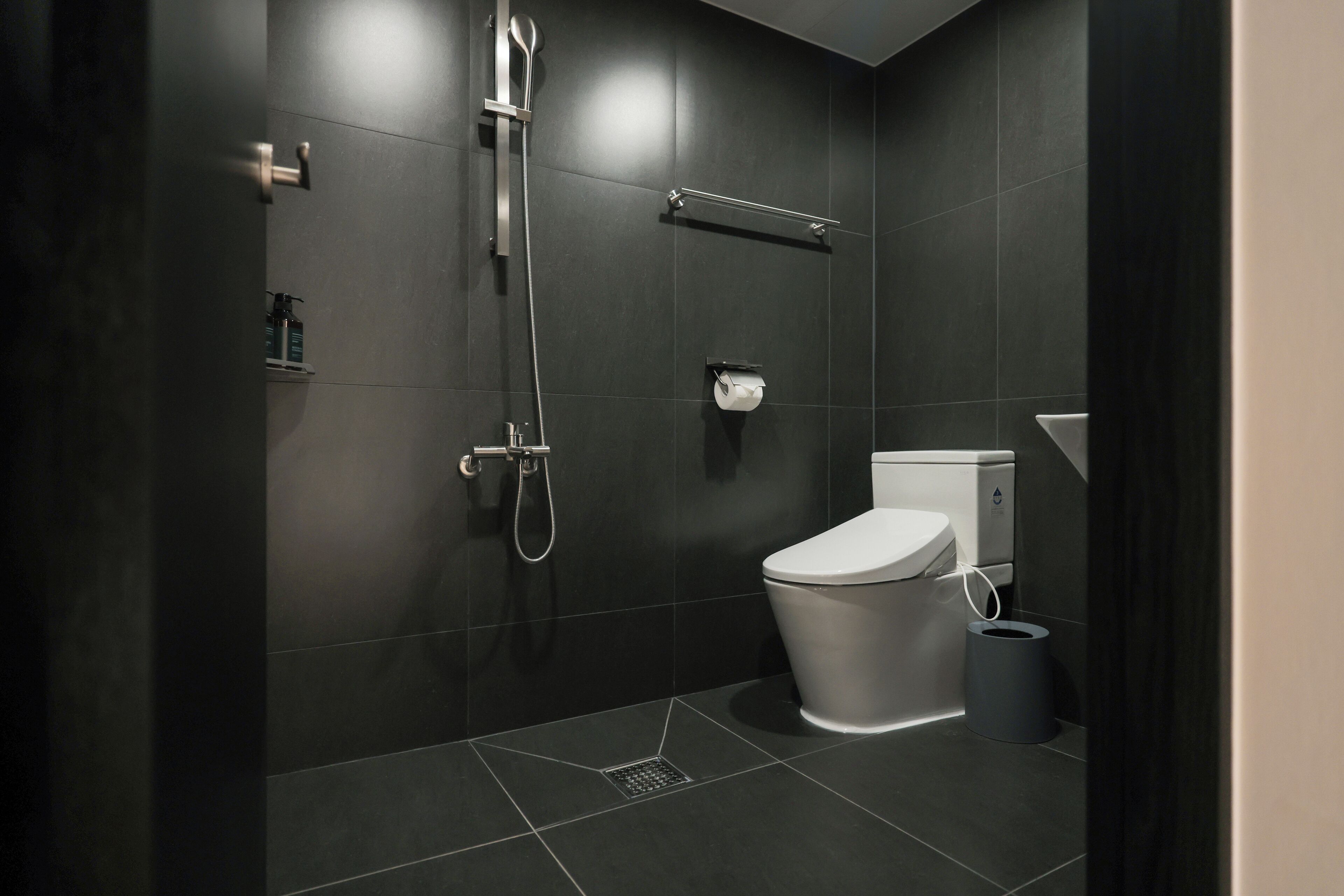 Een moderne badkamer met donkere tegels, een toilet, een douche en een prullenbak.