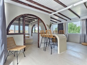 Una habitación con techo alto, vigas de madera, cama, una mesa de comedor con sillas y grandes ventanales con vistas a zonas verdes.