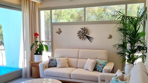 A modern living room with a beige sofa, round coffee table, and a large window with a view of greenery.