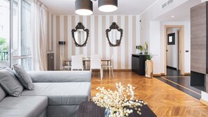 A modern living room with a grey sofa, a wooden coffee table with a floral arrangement, a dining area with a white table and chairs, and a kitchen area with a black countertop and a potted plant. - Easylife - Splendid flat with balcony Central FS (Milano)