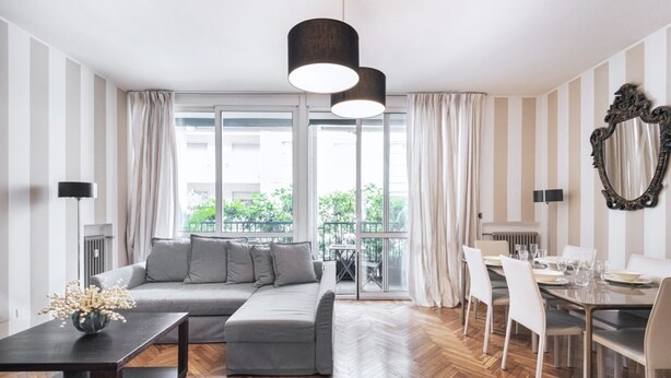 A modern living room with a grey sectional sofa, a dining table with chairs, and a large window with curtains. - Easylife - Splendid flat with balcony Central FS (Milano)