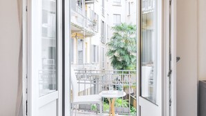 Eine offene Tür zu einem Balkon mit weißem Stuhl und Tisch, einer Palme und Blick auf ein mehrstöckiges Gebäude mit Balkonen.