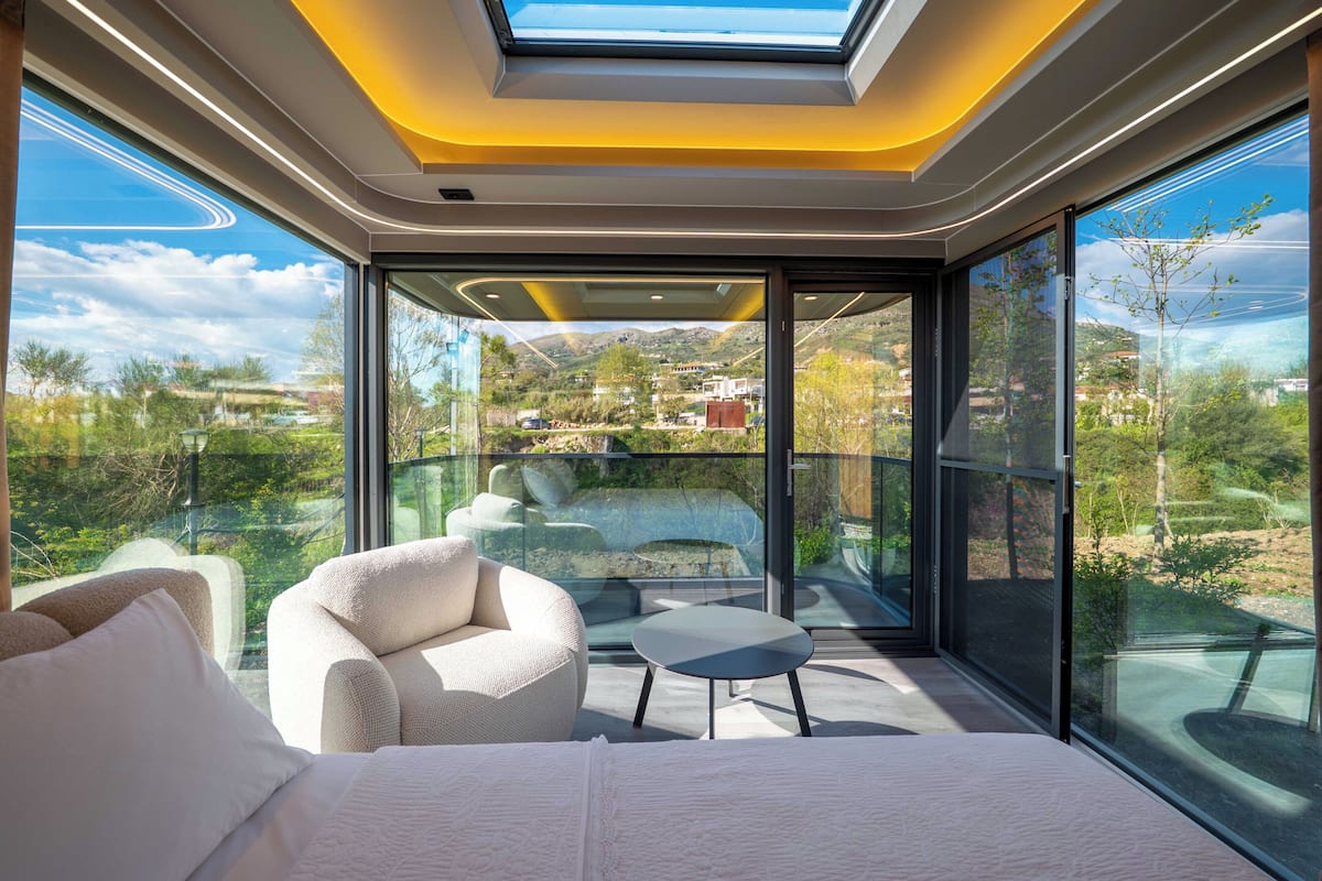 Una habitación moderna con cama, sofá, una mesita y un muro de cristal con vistas panorámicas.