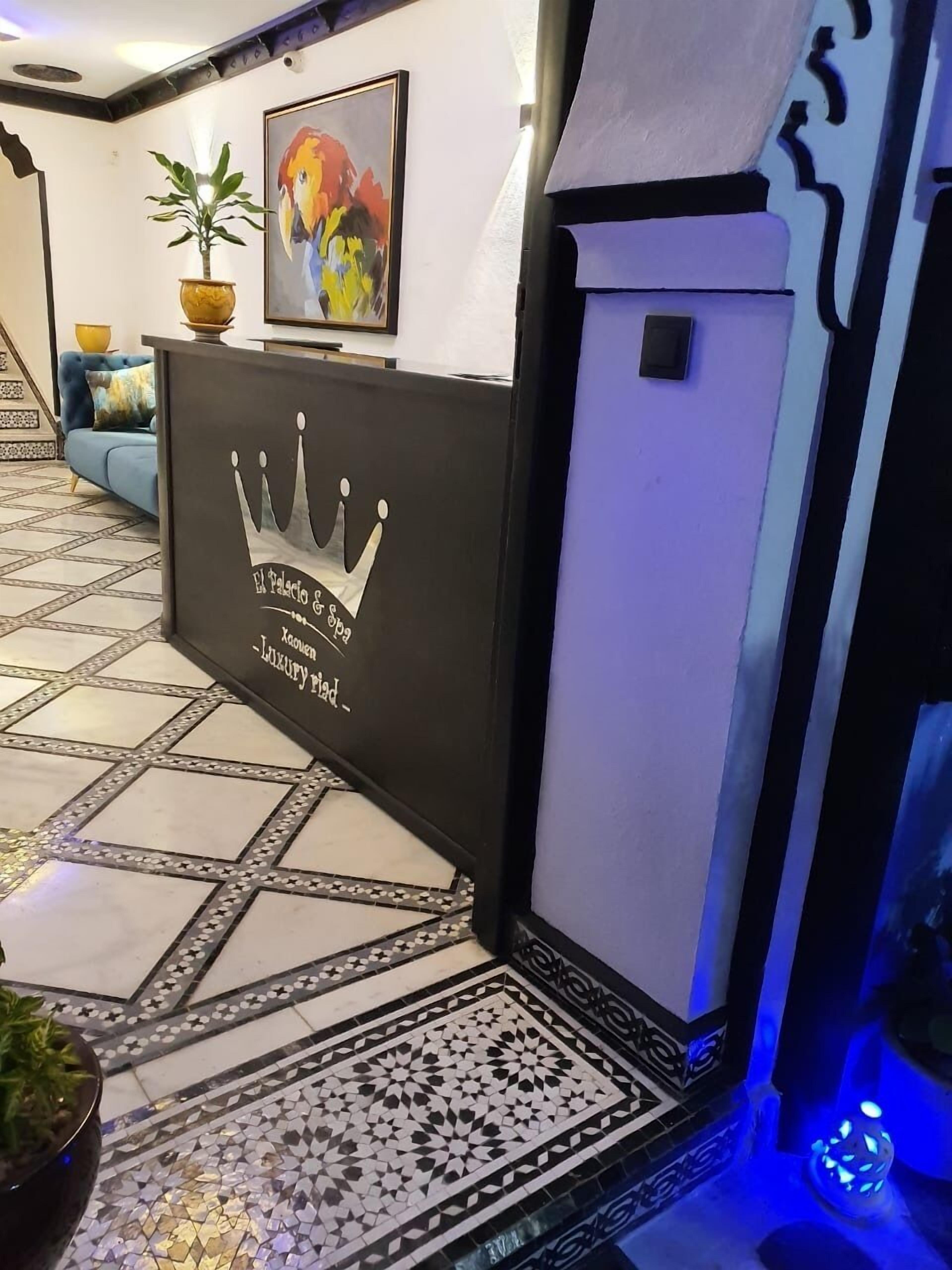 Un espace d’accueil agrémenté d’une œuvre d’art encadrée, d’une plante en pot et d’un comptoir de réception orné d’un logo en forme de couronne.