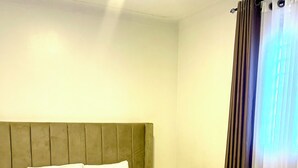 A neatly made bed with a headboard, a bedside table, and a window with curtains. - Cozy amazing one bedroom unit Mariam  (Kampala)