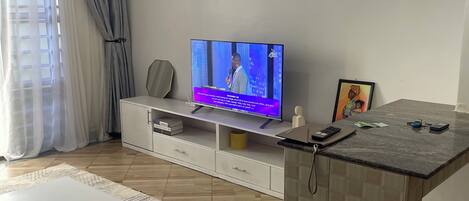 A modern living room with a flat-screen TV, a white coffee table, a patterned rug, and a kitchen island with a tiled backsplash.