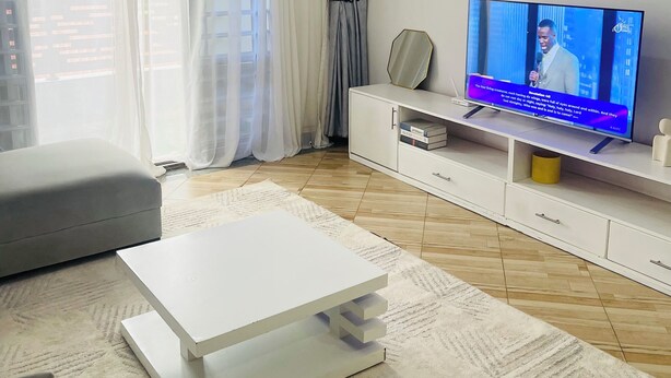 A modern living room with a flat-screen TV, a white entertainment unit, a grey sofa, and a patterned rug. - Cozy amazing one bedroom unit Mariam  (Kampala)