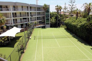 En tennisbane omgivet af en høj bygning med altaner, palmer og en hæk.