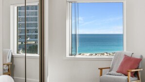 A room with a large window offering a view of the ocean and a building across the street.