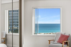 A room with a large window offering a view of the ocean and a building across the street.