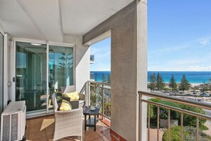 A balcony with a glass door leading to an indoor area, a wicker chair with cushions, a small table, and a view of the ocean and beach.