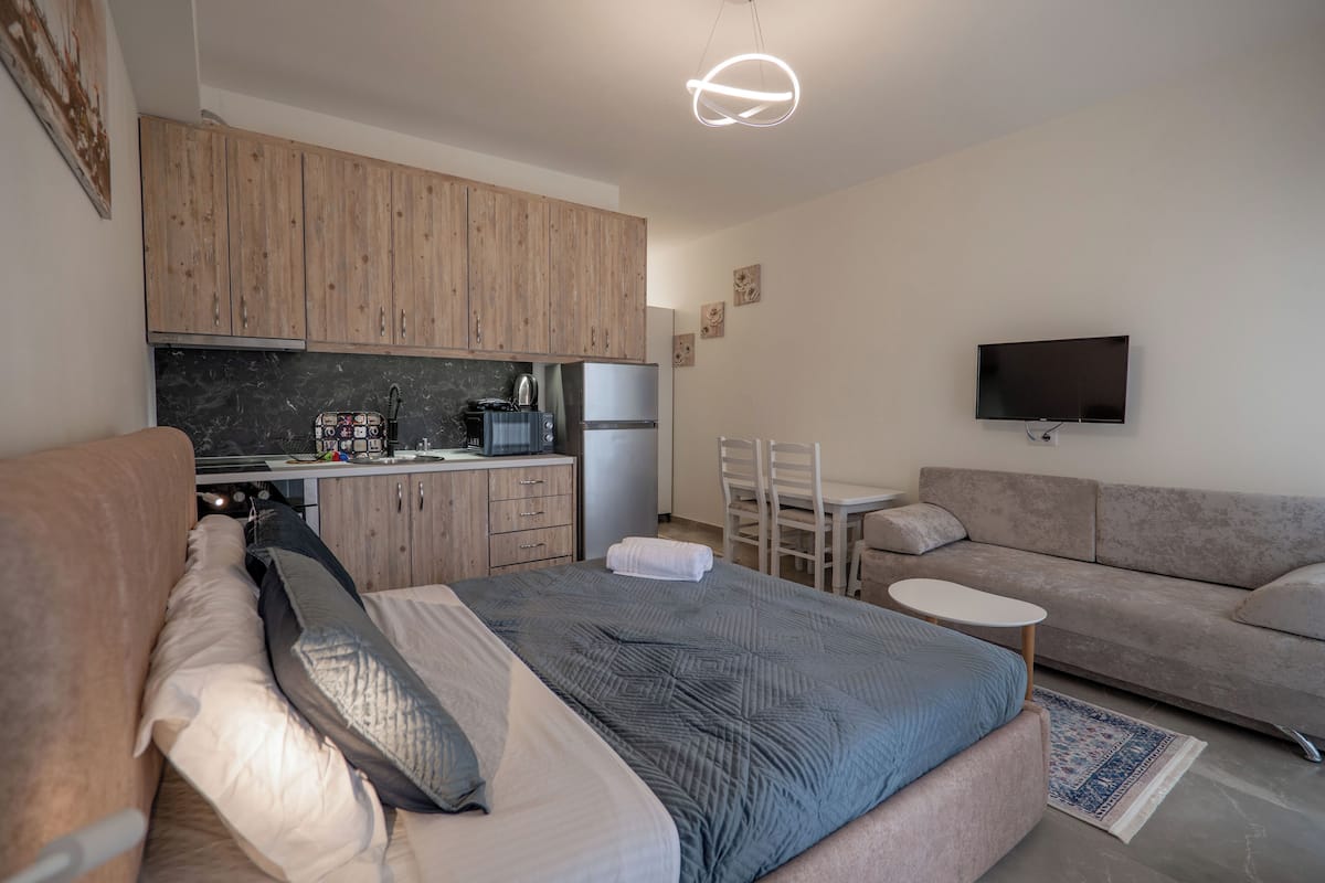 A modern bedroom with a large bed, a small table, a sofa, and a kitchen area with wooden cabinets and a flat-screen TV.