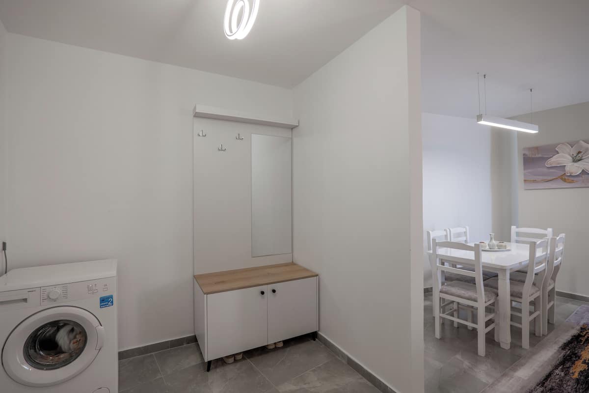 A compact living space with a washing machine, a wooden cabinet, a dining table with chairs, and a ceiling light fixture.