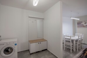 A compact living space with a washing machine, a wooden cabinet, a dining table with chairs, and a ceiling light fixture. - Esel Apartments (Sarandë)