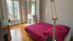 Une chambre avec un plancher en bois, un lit recouvert d’une couverture rouge, une table de chevet blanche, un miroir et une lampe sur pied.