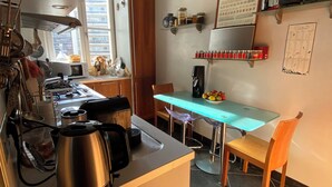 Une cuisine compacte équipée d’une cuisinière à gaz, d’une bouilloire et de divers ustensiles. Elle comprend un coin repas avec une table et des chaises, ainsi qu’une fenêtre qui laisse entrer la lumière naturelle.