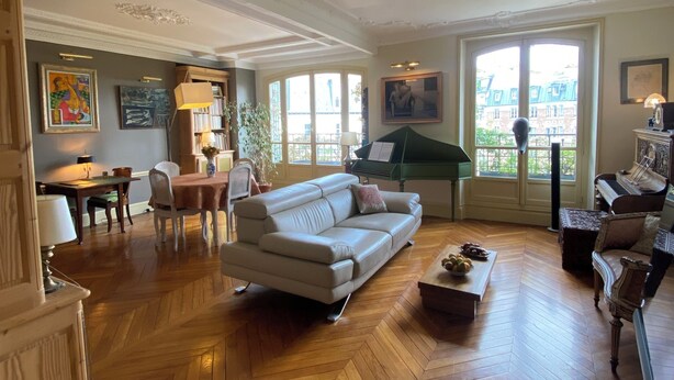 Un vaste salon doté d’un parquet en point de Hongrie, d’un piano, d’un coin repas et offrant une vue dégagée grâce à de grandes fenêtres.