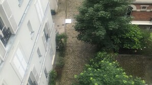 Une ruelle étroite pavée, bordée de bâtiments aux façades blanches et aux volets fermés, une voiture rouge garée sur le côté droit, et de luxuriants arbres verdoyants.