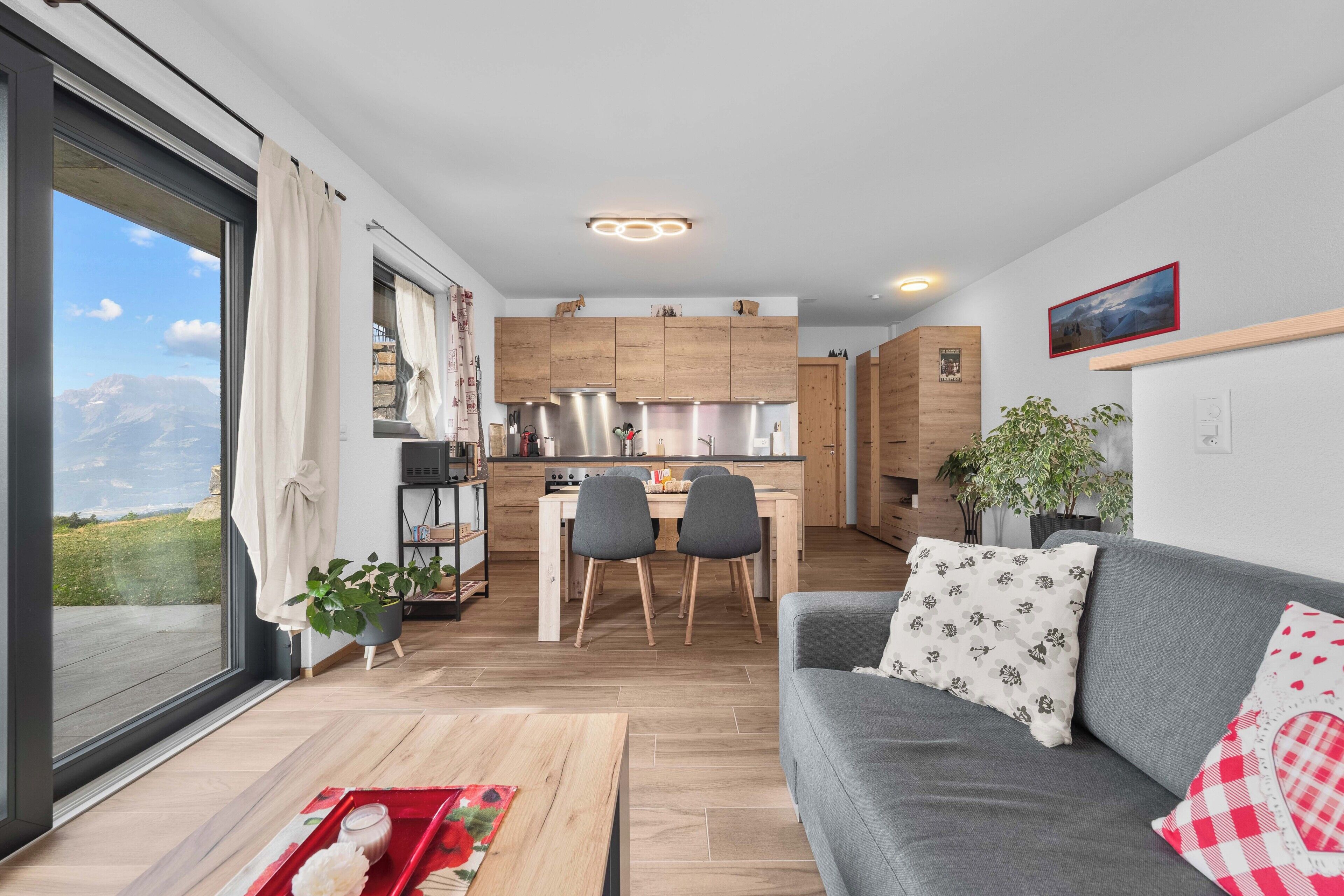 Una sala de estar moderna con un sofá gris, un comedor con mesa y sillas, y una cocina con gabinetes de madera.