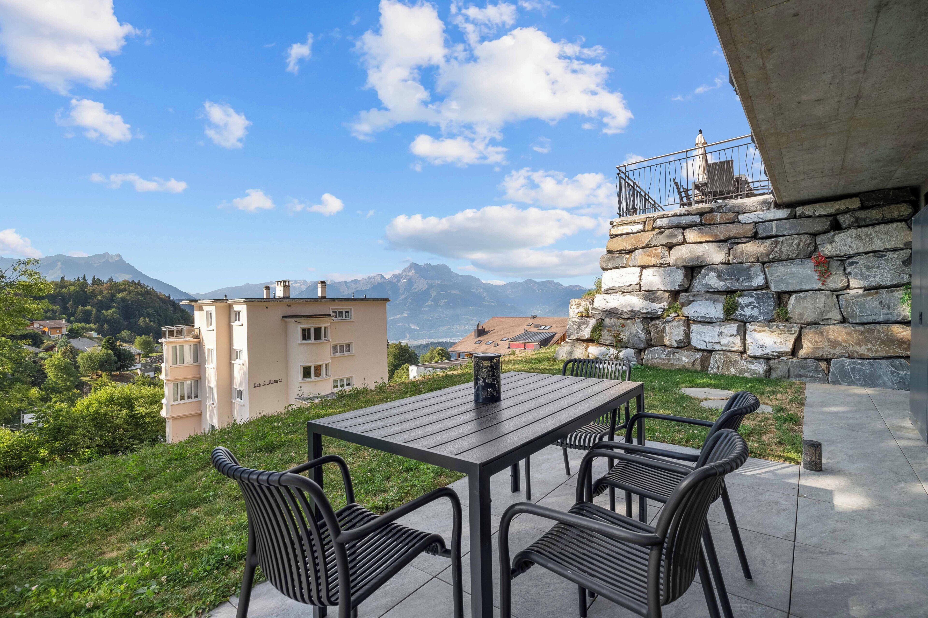An outdoor patio with a table and chairs, overlooking a mountainous landscape and a residential area.