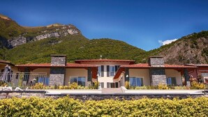 Une maison moderne à deux étages, dotée d’une façade en pierre, de grandes fenêtres et d’un toit recouvert de tuiles rouges, qui se dresse sur un fond de montagnes.