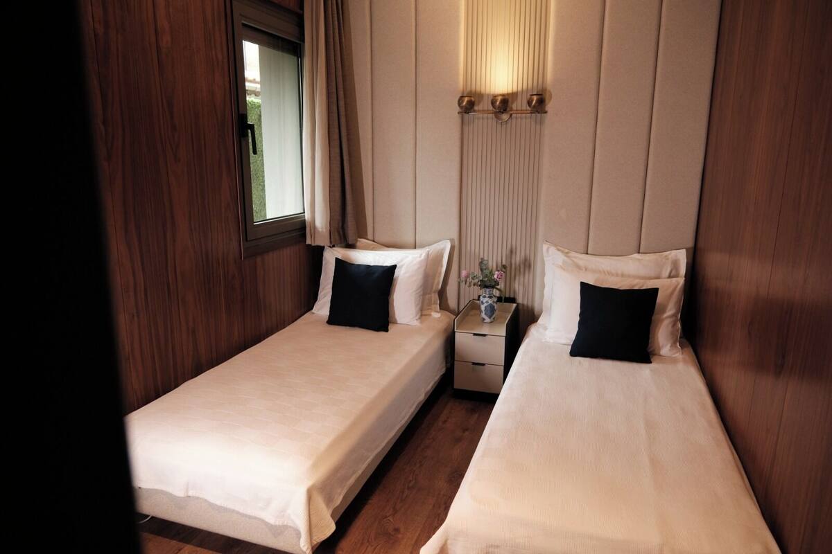 A small, cozy room with two single beds, a nightstand with a vase, and a window with curtains.