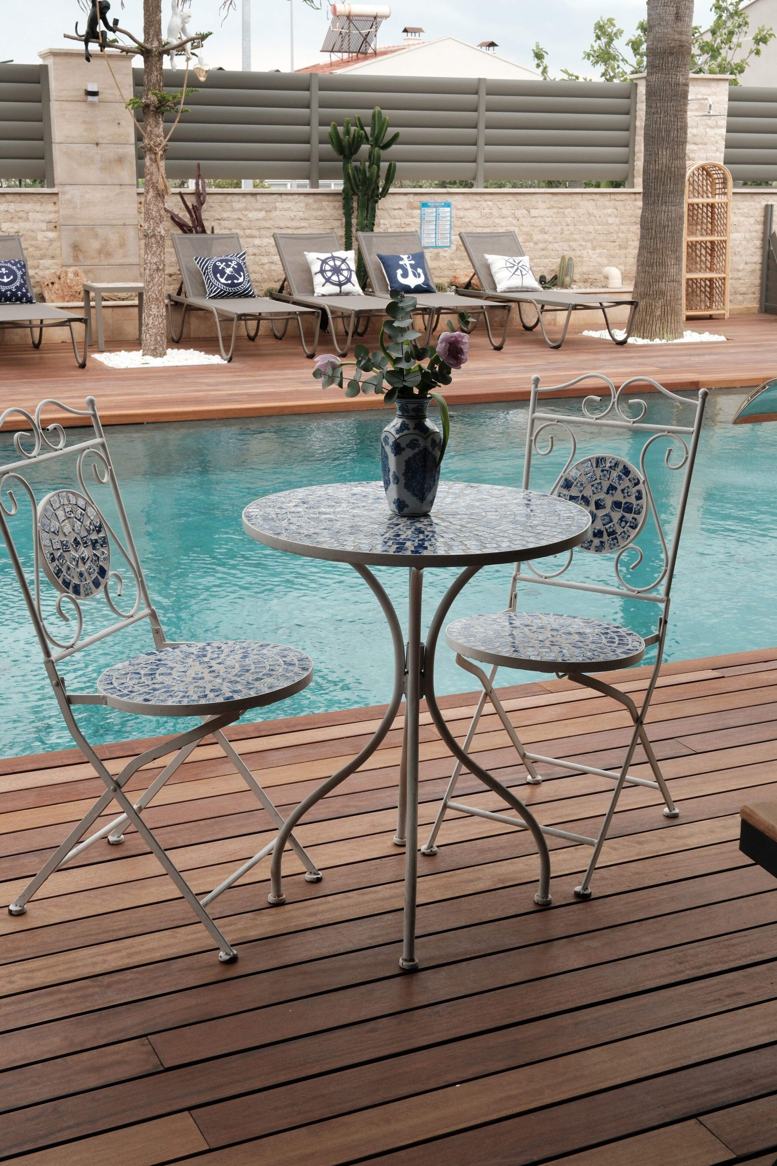 A poolside area with a table and chairs, a vase with flowers, and lounge chairs with cushions.