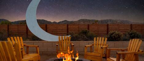 A nighttime outdoor seating area with a fire pit and wooden chairs, a wooden fence, and a backdrop of a starry sky with a crescent moon.